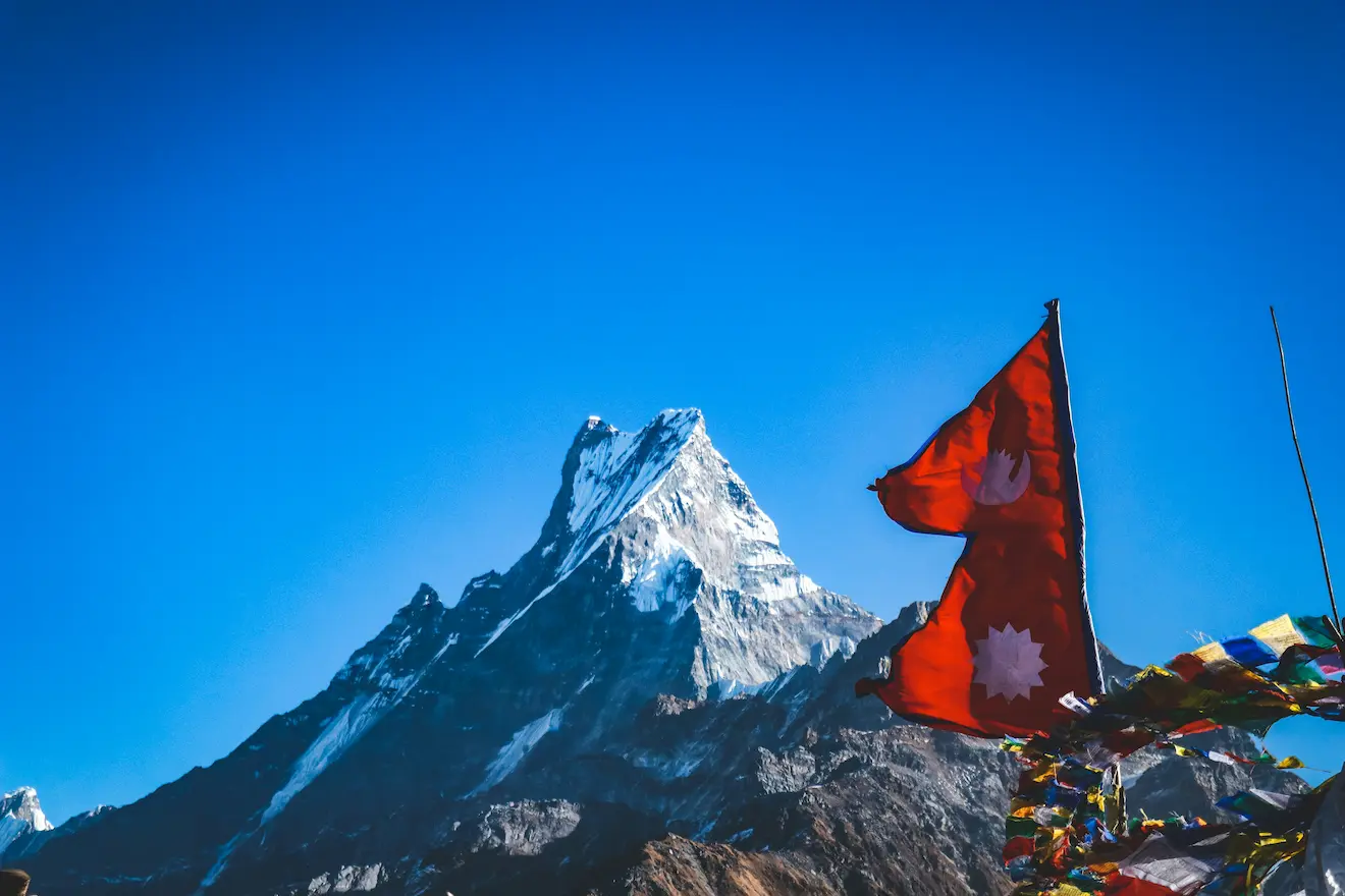 Machhapuchhre, the Fishtail mountain near Pokhara, under a clear blue sky, with the Nepali flag and prayer flags in the foreground.