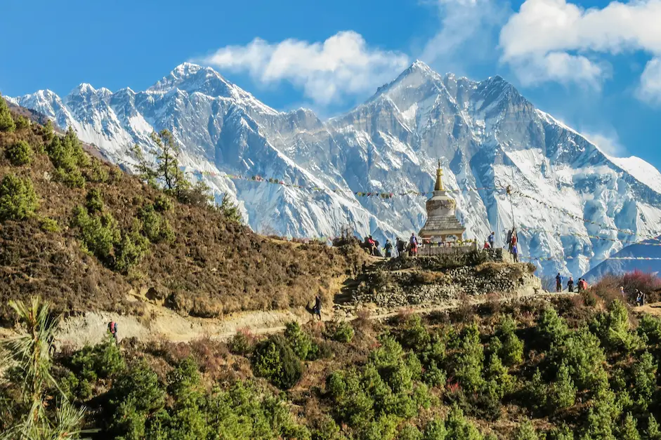 Trekkers passing a white stupa on the trail near Namche Bazaar, with the snow-covered Everest massif rising behind.